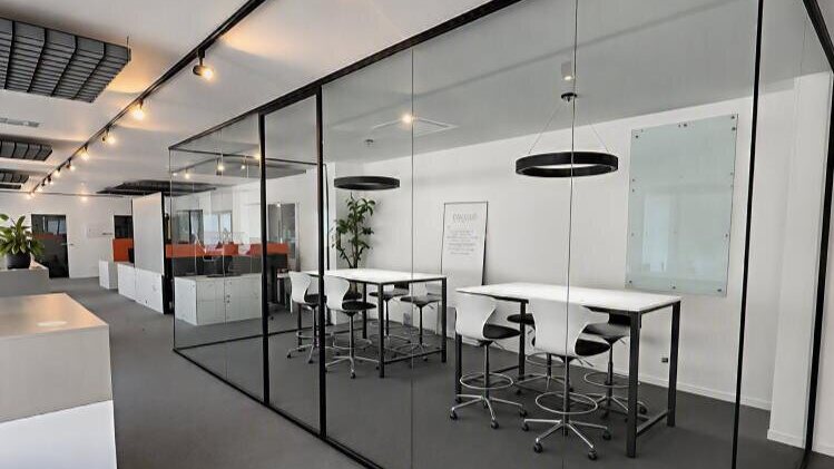 Open-Space-Bürofläche mit schwarz gerahmten Glasboxen, Stehtischen mit Barhockern und Ringleuchten.