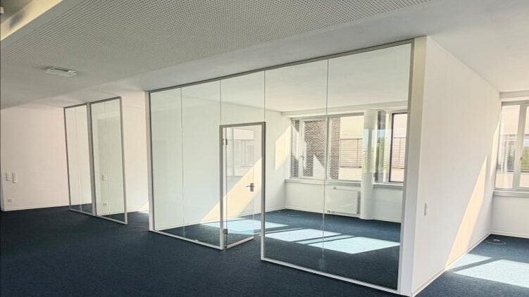 Neu erstellte Bürozellen aus Glastrennwänden vor der Möblierung, mit dunkelblauem Teppichboden und weißer Akustikdecke.